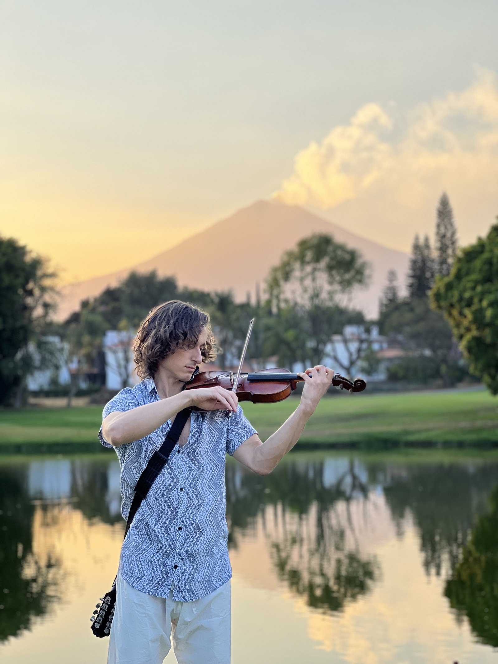Samuel Wexler playing violin at dusk beside a lake in Atlixco, with the Popocatepetl volcano behind him.