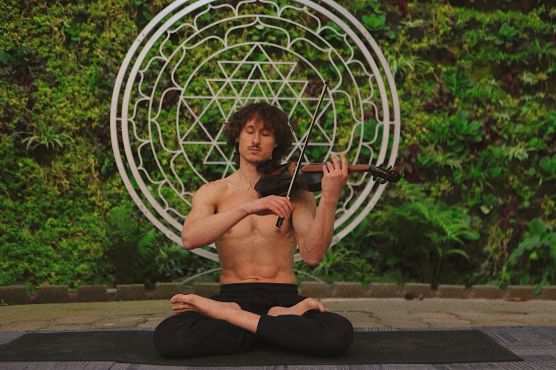 Samuel Wexler seated in lotus with a violin, in front of a Sri Yantra sculpture on a living wall.
